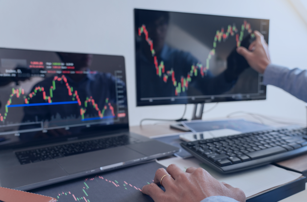 Laptop i ekran komputera wyświetlające wykres rynku akcji. Czyjaś ręka wodzi po nim palcem.