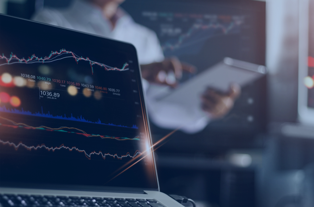 Zbliżenie na ekran laptopa wyświetlającym wykres giełdy. W tle inwestor używający tableta.