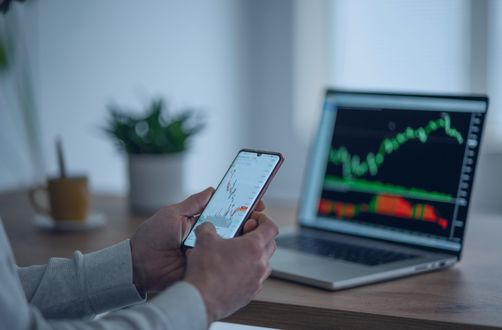 Zbliżenie na ręce, w których trzymany jest telefon komórkowy z wykresem akcji. W tle laptop wyświetlający podobny wykres.