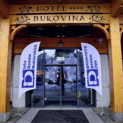 Wejście do Hotelu Bukovina z drewnianą architekturą w stylu góralskim; nad drzwiami szyld „Hotel Bukovina”, przed wejściem dwie flagi Izby Domów Maklerskich.